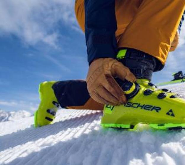 Sciatore allaccia scarponi gialli su pista innevata.